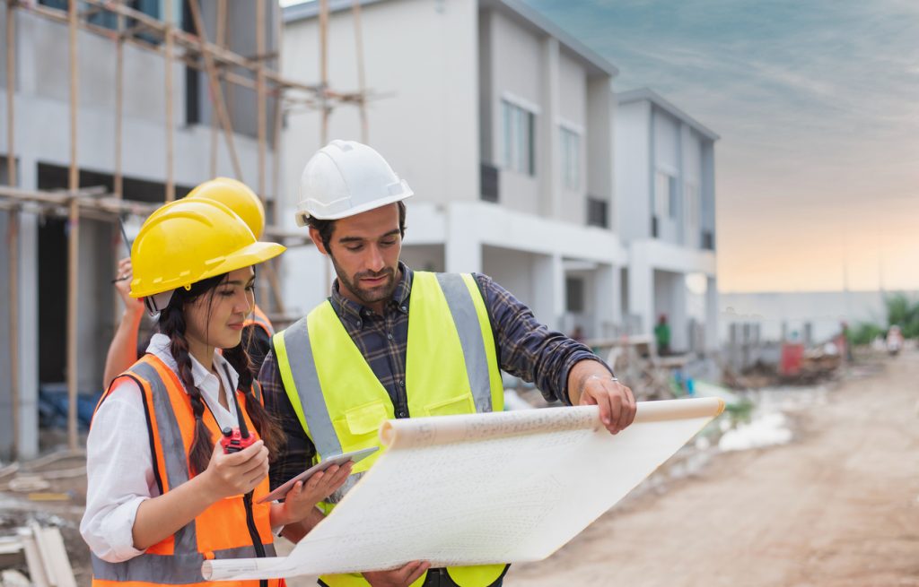 Caucasian males and Asian female builders, architector and engineers with draft plan of building and laptop computer talking on constructing site